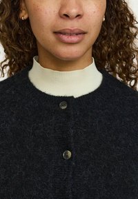 Czarny wełniany kardigan z kremowym prążkowanym kołnierzem, zapięciem na guziki z przodu oraz miękką, puszystą fakturą. Wyraziste guziki dodają szczegółów.