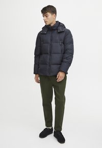 Marineblå pufferjakke med hette, glidelås foran og sidelommer. Båret med olivengrønne bukser og svarte sko. Sletten, quiltet tekstur.