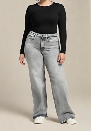 Vrouw draagt een zwart, nauwsluitend top met lange mouwen, lichtgrijze, wijde, hooggetailleerde jeans en witte sneakers, staand tegen een beige achtergrond.