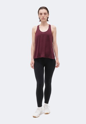 Giovane persona in piedi con canottiera bordeaux senza maniche, leggings neri, scarpe da ginnastica bianche e calzini grigio chiaro su sfondo semplice.