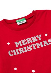 Camiseta de algodón roja con el texto "FELIZ NAVIDAD" en purpurina plateada. Cuello redondo con detalles de lunares blancos en la tela.