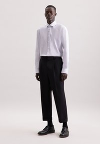 Camicia bianca a maniche lunghe con colletto strutturato, abbinata a pantaloni neri plissettati e scarpe nere con lacci, su uno sfondo neutro.