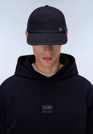 Mann trägt eine schwarze Kappe mit einem kleinen norwegischen Flaggenemblem und einen schwarzen Kapuzenpullover mit einem kaum sichtbaren rechteckigen Logo auf der Brust.
