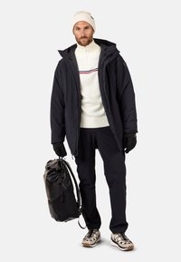 Giubbotto imbottito nero, maglione bianco con righe orizzontali, guanti neri, pantaloni blu, porta uno zaino nero e indossa un berretto di maglia leggera.