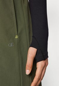 Green athletic wear with a close-up of a black long-sleeve shirt sleeve. Features a thumbhole and visible stitching details.