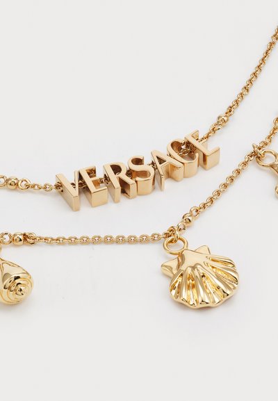 Gold necklace featuring "VERSACE" letters with a polished finish, accented by a rose and shell charm, on a delicate chain.