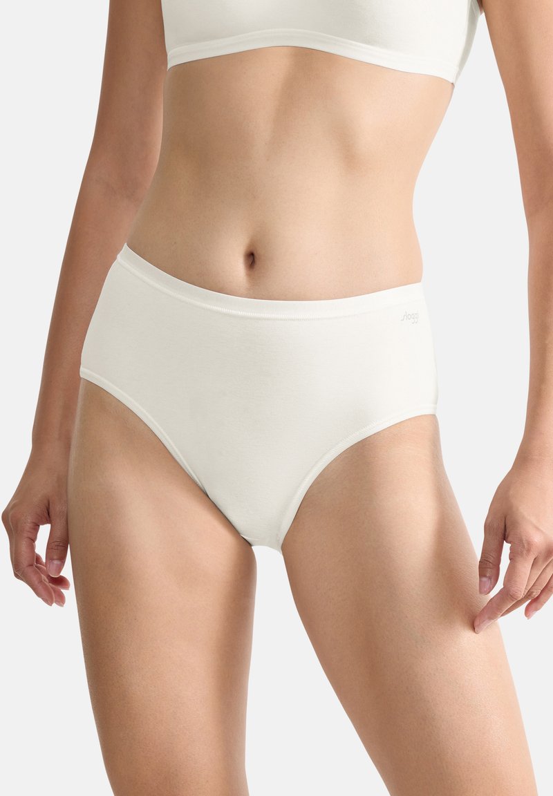 Femme portant une lingerie blanche en coton taille haute et un soutien-gorge assorti, debout avec les bras détendus devant un fond gris clair.