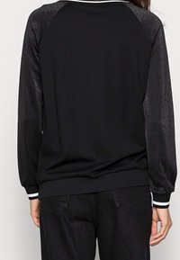 Osoba ubrana w czarny sweter z długim rękawem, z błyszczącymi czarnymi rękawami oraz białymi paskami na mankietach i kołnierzu, w połączeniu z czarnymi spodniami.