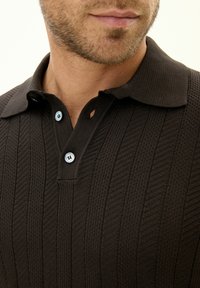 Brun texturerad polo-shirt med ett ribbat mönster, krage och tvåfärgade knappar. Närbild med fokus på halsringningen och tygdetaljerna.