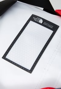 White bag featuring a transparent hexagonal-patterned ID window, framed in black with stitching accents, and a black strap and red detail.