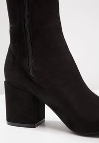 Bottine en daim noir avec un talon carré bloc, une fermeture éclair latérale, et une texture lisse, conçue pour une silhouette ajustée.