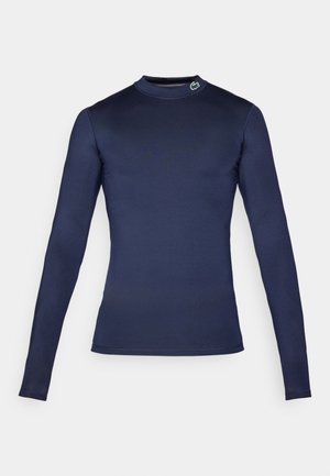 Maglietta a maniche lunghe di colore blu navy realizzata in tessuto liscio ed elasticizzato. Presenta un collo alto e un piccolo dettaglio del logo verde sul colletto.