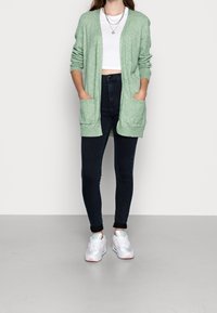 Světlé zelené pletené kardigan s otevřeným předním dílem, dlouhými rukávy a dvěma předními kapsami, kombinovaný s tmavými džínami a bílými teniskami.