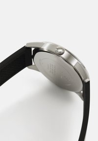 Boîtier de montre en acier inoxydable argenté de forme ronde, bracelet en caoutchouc noir, accents texturés et couronne texturée sur le côté.