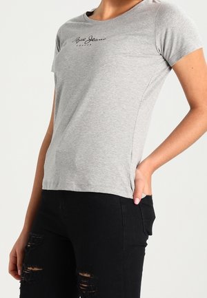 Kvinde iført en tætsiddende grå kortærmet t-shirt med "Pepe Jeans London" logo og sorte revnede jeans, den ene hånd i lommen.