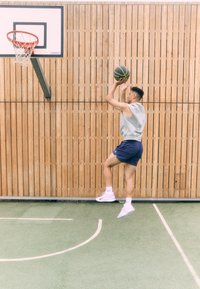 A person jumping to shoot a basketball towards a hoop. They wear a gray sleeveless top, blue shorts, and white athletic shoes on a green court.