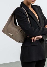 Braune Stoffhandtasche mit einem lässigen Design, das eine dezente Textur und goldene Hardware-Details zeigt. Getragen über einem schwarzen Blazer.