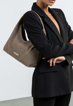Bruine stoffen handtas met een slouchy ontwerp, die een subtiele textuur en gouden hardwareaccenten toont. Draagover een zwarte blazer.