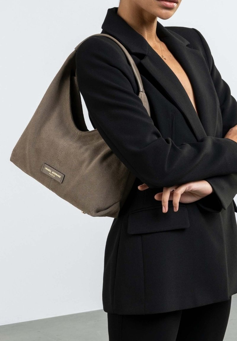 Braune Stoffhandtasche mit einem lässigen Design, das eine dezente Textur und goldene Hardware-Details zeigt. Getragen über einem schwarzen Blazer.