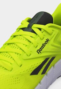 Φωτεινό κίτρινο αθλητικό παπούτσι Reebok με μαύρο λογότυπο, κορδόνια και αναπνεύσιμο πλέγμα στο επάνω μέρος, που δείχνει λεπτομέρειες από την πλευρά και τη γλώσσα του παπουτσιού.
