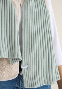Lys grøn ribbet tørklæde lavet af blødt strikmateriale, med et mærke med "CECIL." Tørklædet vises mod en baggrund af en beige vest.