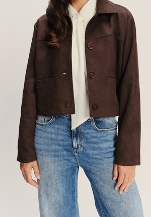 Vrouw die een donkerbruin cropped jasje met grote knopen draagt over een witte blouse met strik, gecombineerd met lichtblauwe spijkerbroek.