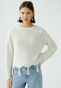Weißer gestrickter Pullover mit einem Zickzackmuster, mit abgerundetem Saum und fransigen Akzenten. Kombiniert mit hellblauen Jeans.