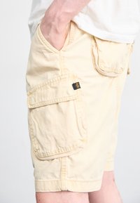 Shorts cargo de culoare galben deschis, cu două buzunare laterale, un buzunar la spate și un etichetă din material pe partea superioară a buzunarului din dreapta.