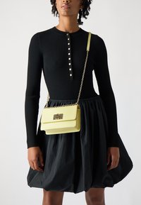 Top lungo a maniche rigide nero con bottoni dorati, abbinato a una gonna nera voluminosa e una borsa in pelle gialla con tracolla a catena.