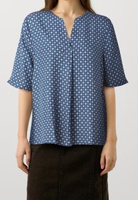 Blusa blu a fantasia con maniche corte, scollo a V, caratterizzata da forme geometriche in bianco e rosso. Tessuto leggero con una texture liscia.