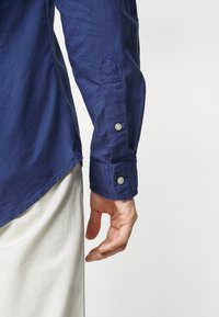 Henkilön oikea käsi roikkuu alas, hänellä on päällään pitkähihainen sininen paita, jonka hihansuu on napitettu, ja vaaleat housut, taustana yksivärinen pinta.