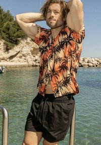 Chemise à manches courtes, orange et crème, avec imprimé palmier, assortie à un short de bain noir, debout près de l'eau.