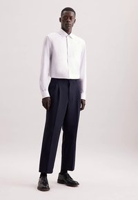 Wit shirt met lange mouwen en een borstzak, gecombineerd met marineblauwe geplooide broek. Zwarte leren schoenen maken de look af. Soepele, op maat gemaakte pasvorm.