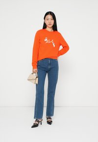 Felpa arancione con grafica bianca, jeans blu a gamba dritta, scarpe con tacco nere con decorazioni e una piccola borsa beige.