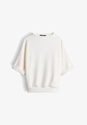 Witte blouse met korte mouwen, opgerolde manchetten, ronde halslijn en elastische taillezoom tegen een witte achtergrond.