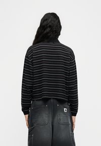 Άτομο με μακριά, κυματιστά μαλλιά που φοράει μαύρο πουκάμισο με μακριά μανίκια και λεπτές λευκές ρίγες, καθώς και σκουρόχρωμο γκρι τζιν με μεγάλες πίσω τσέπες.