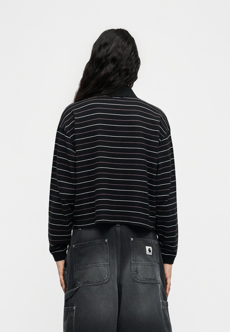 Persoon met lang golvend haar, gekleed in een zwart shirt met lange mouwen met dunne witte strepen en donkergrijze jeans met grote achterzakken.