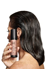 Rehabilitujący nawilżacz do suchych włosów GHD w matowej czarnej butelce ze złotymi akcentami, 256 mL. Użytkownik trzyma butelkę, a widać błyszczące, mokre włosy.