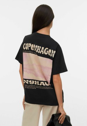Person med langt mørkt hår iført sort T-shirt med stor tekst 'Copenhagen Nyhavn' og en lyserød bysilhuet på ryggen, der holder en sort taske.