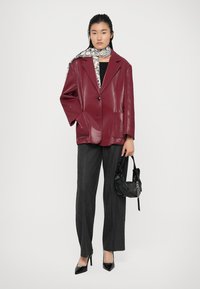 Blazer in finta pelle color bordeaux con rever a alette, abbinato a pantaloni a righe. Accessoriato con una borsa perforata nera e una sciarpa stampata.