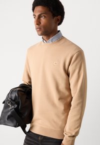 Sweatshirt en coton beige avec des poignets et un col côtelés. Présente un patch logo texturé sur la poitrine. Sac en cuir noir avec une sangle.