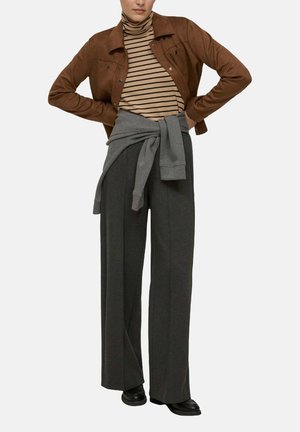 Giacca di camoscio marrone, dolcevita a righe, pantaloni grigi a gamba larga con felpa grigia annodata in vita e scarpe nere.