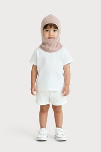 Egy kisgyerek világos rózsaszín balaklávat, fehér pólót, fehér sortot és fehér sportcipőt visel, egy sima háttér előtt áll.