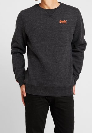 Mężczyzna ubrany w ciemnoszarą bluzę z okrągłym dekoltem z małym pomarańczowym logo "SuperDry" na górnej lewej części piersi oraz czarne spodnie, stoi z luźno opuszczonymi rękami.