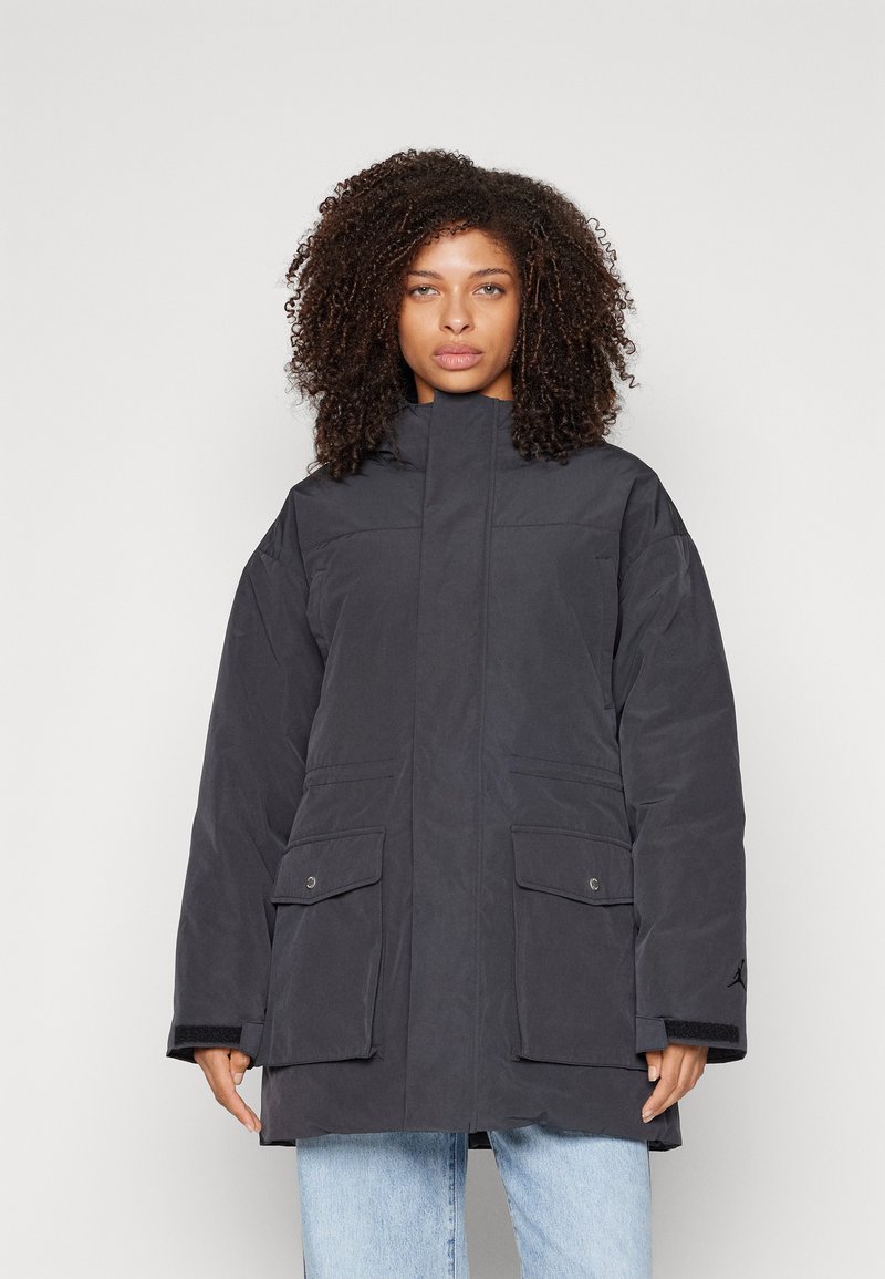 Parka noire rembourrée avec des manches longues, un col montant et deux poches avant. Présente un tissu texturé et une marque subtile sur la manche.