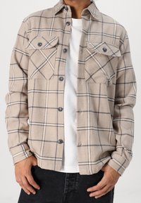 Beige geruite shirt met twee borstzakken, ronde kraag en knoopsluiting. Zachte textuur met donkergrijze accenten en contrasterende witte lijnen.