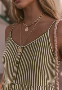 Grün-weiß gestreiftes Kleid mit Spaghettiträgern, das eine Knopfleiste und einen gestuften Saum hat. Goldene, geschichtete Halsketten mit runden Anhängern.