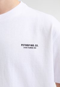 Bílý bavlněný tričko s kulatým výstřihem a krátkými rukávy. Na levé straně hrudi je černý text "PETROLIND CO. 31224 FLORIDA USA".