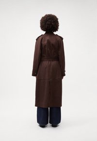 Cappotto trench marrone con mostrine sulle spalle, vita cinchata e design lungo, abbinato a jeans denim a gamba larga. Tessuto di facile consistenza.