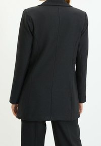 Svart skräddarsydd kavaj med en slät textur, långa ärmar och kavajkrage. Har vertikala sömmar och en rak skuren bakdel.